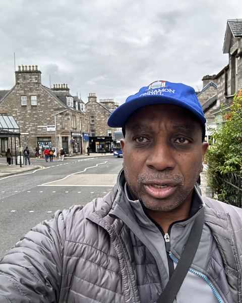 Man in a blue cap and grey jacket taking a selfie on a Scottish high street