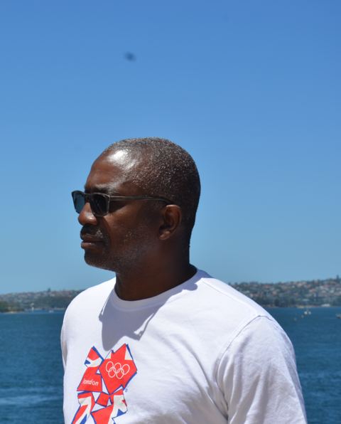 Man in sunglasses and a white T-shirt in profile, with a blue harbour behind him